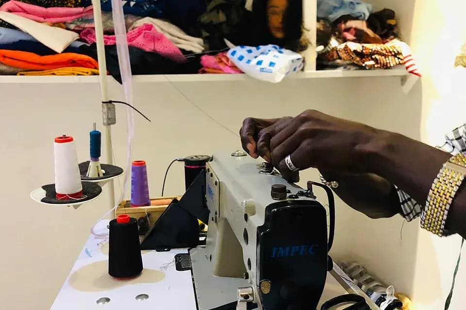 Atelier de couture au Sénégal : les mains d'un tailleur près d'une machine à coudre, avec fils et tissus sur les étagères