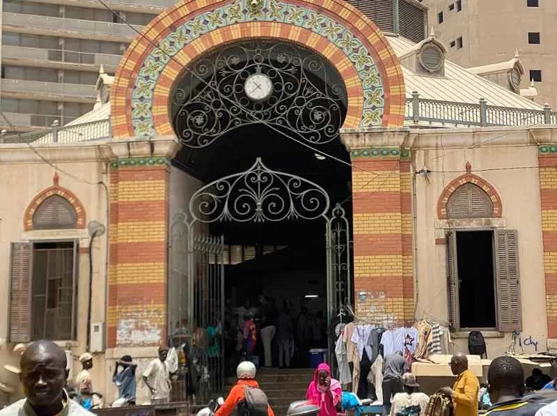Façade en briques jaunes et rouges du marché Kermel à Dakar, avec son grand arc, ses grilles en fer forgé et des passants devant l'entrée