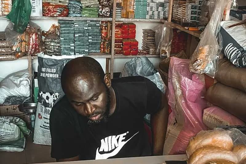 Commerçant derrière le comptoir de sa boutique de quartier, entre sacs de marchandises et étagères de produits