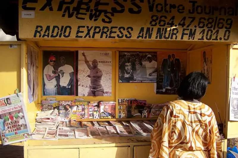 Passante devant un kiosque à journaux de Dakar, quotidiens et magazines exposés sur le comptoir